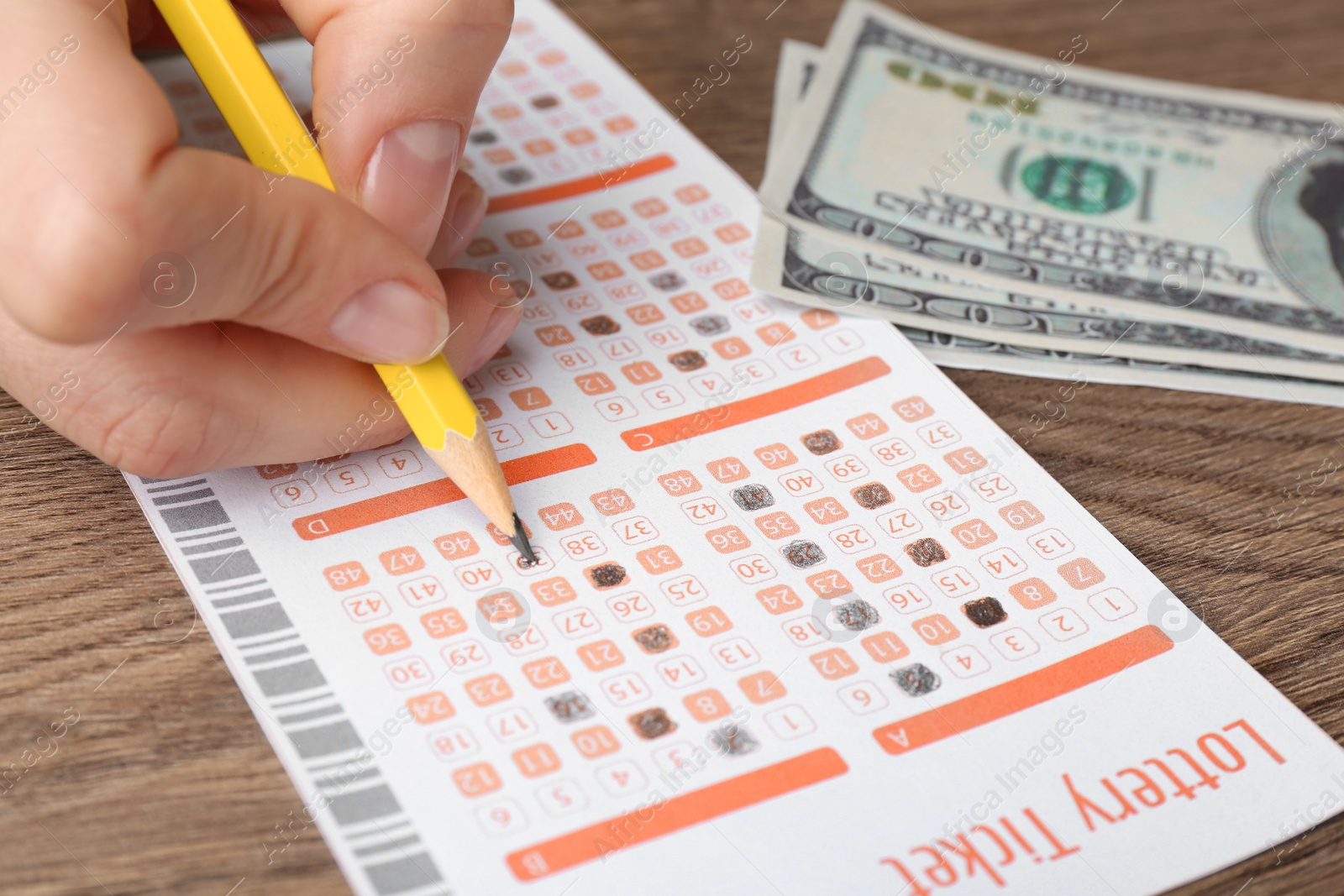 Photo of Woman filling out lottery ticket with pencil and money on wooden table, closeup