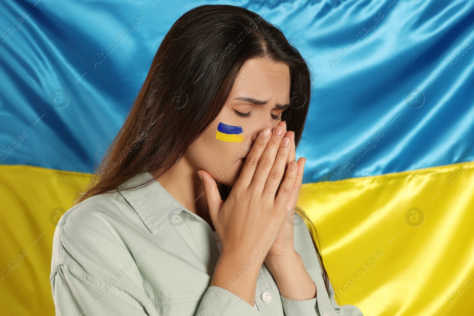 Photo of Sad young woman with clasped hands near Ukrainian flag. Space for text