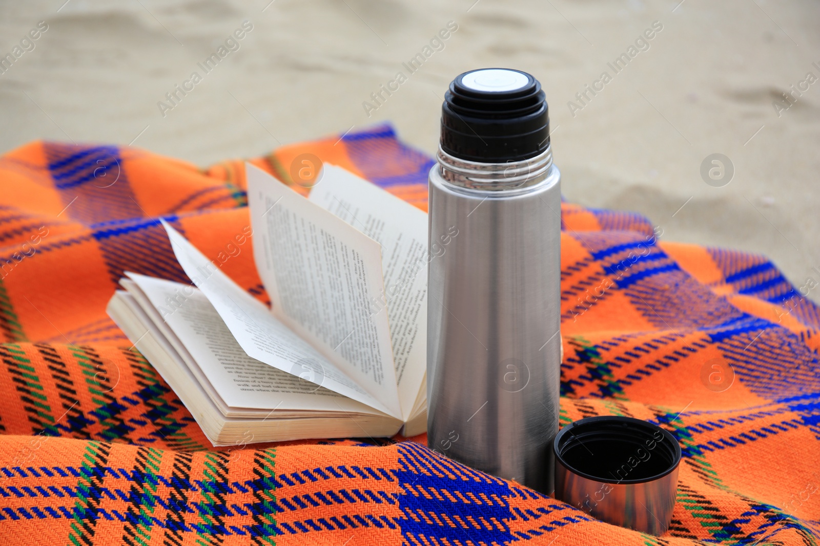 Photo of Metallic thermos with hot drink, open book and plaid on sandy beach, closeup