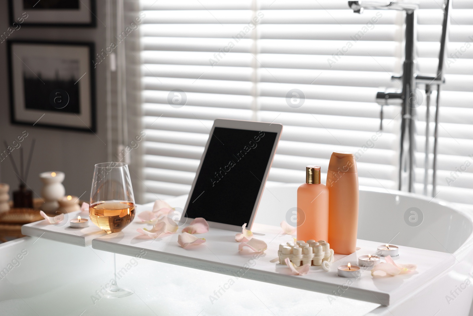 Photo of Wooden tray with tablet, wine, toiletries and flower petals on bathtub in bathroom