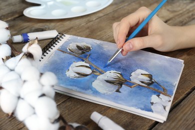 Photo of Woman painting cotton flowers in sketchbook at wooden table, closeup