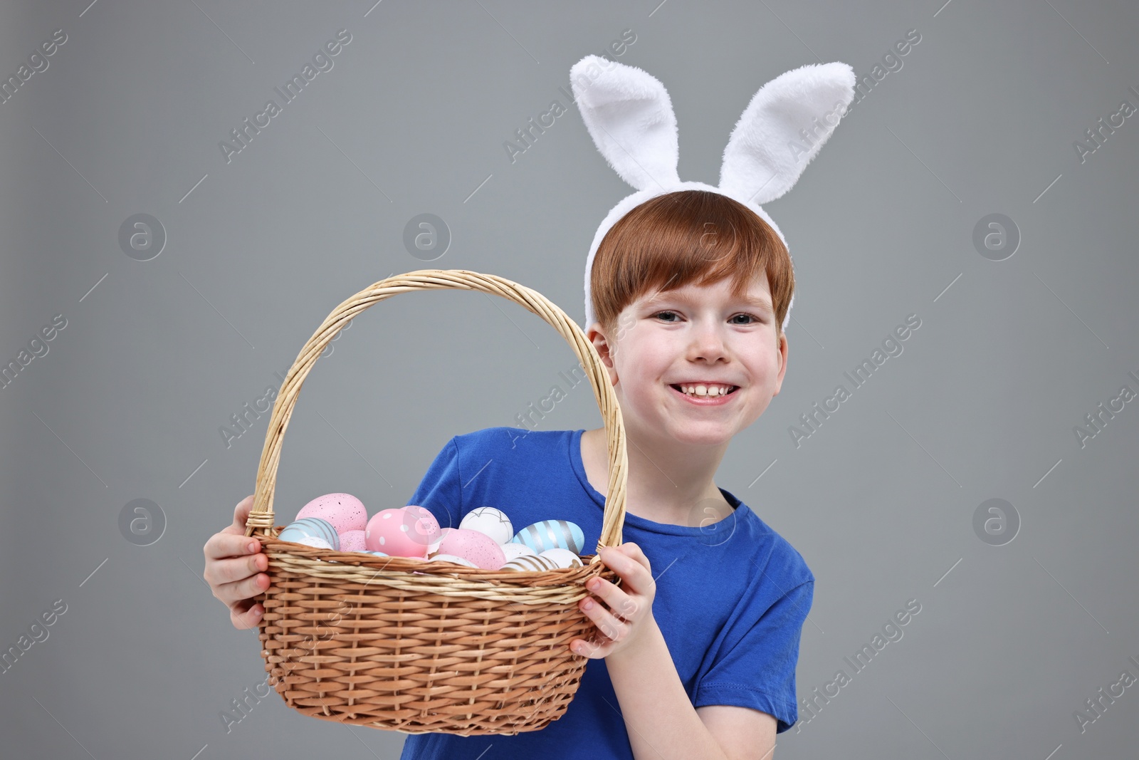 Photo of Easter celebration. Cute little boy with bunny ears and wicker basket full of painted eggs on grey background
