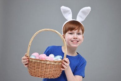 Easter celebration. Cute little boy with bunny ears and wicker basket full of painted eggs on grey background