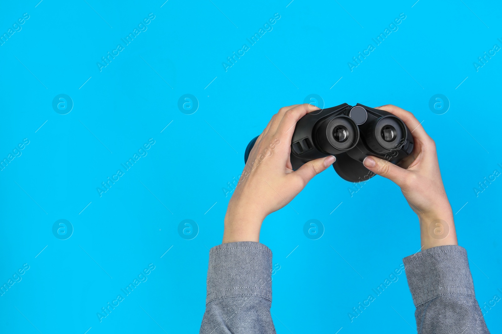 Photo of Woman holding modern binoculars on light blue background, closeup. Space for text