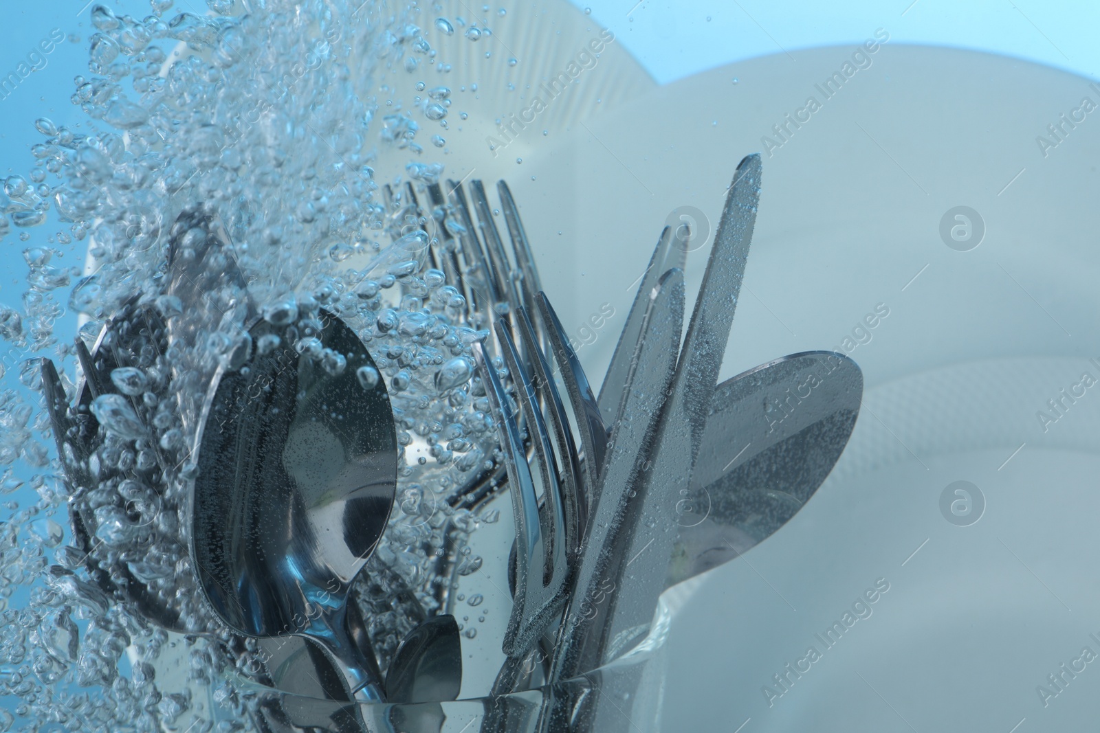 Photo of Washing silver cutlery and plates in water on light blue background, closeup