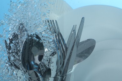 Washing silver cutlery and plates in water on light blue background, closeup