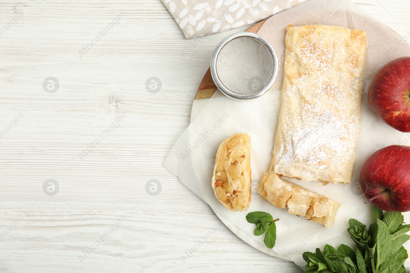 Photo of Delicious strudel, powdered sugar, mint and fresh apples on white wooden table, flat lay. Space for text