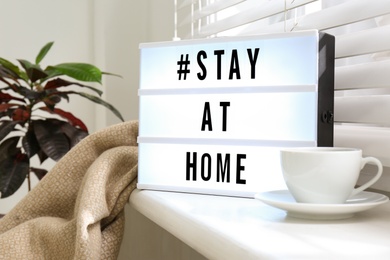 Photo of Cup of drink and lightbox with hashtag STAY AT HOME on window sill indoors. Message to promote self-isolation during COVID‑19 pandemic