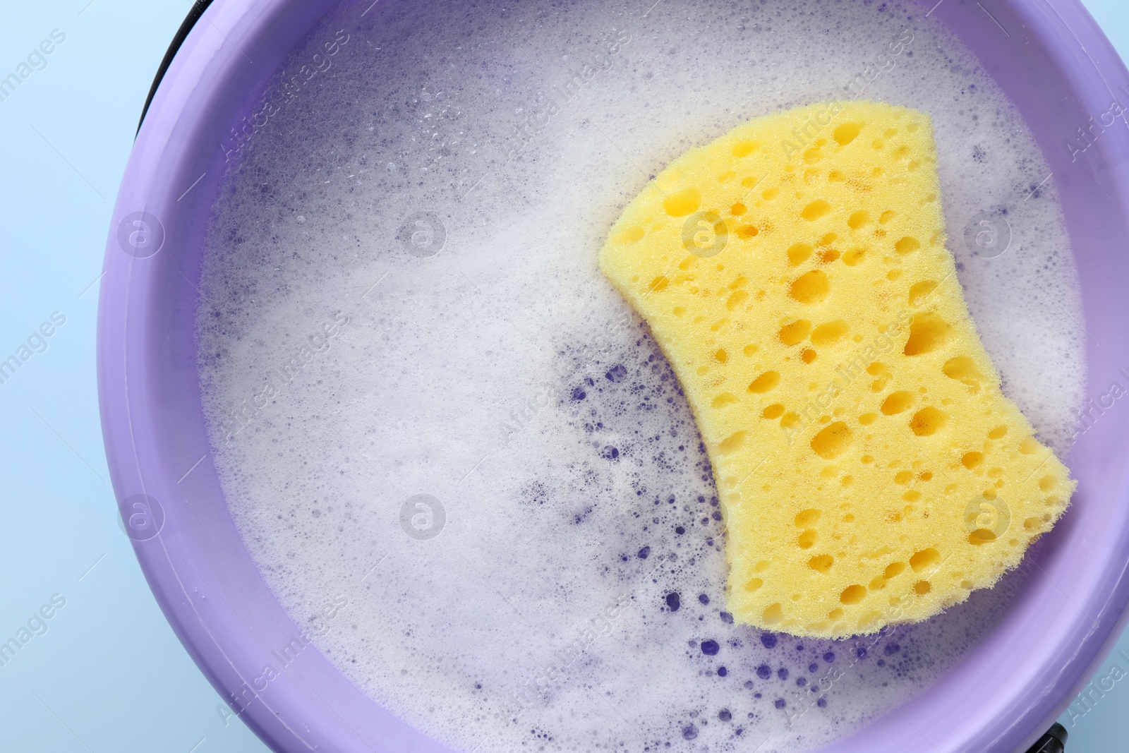 Photo of Bucket with foam and sponge on light blue background, top view