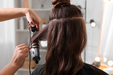 Hairdresser using curling hair iron while working with woman in salon, closeup