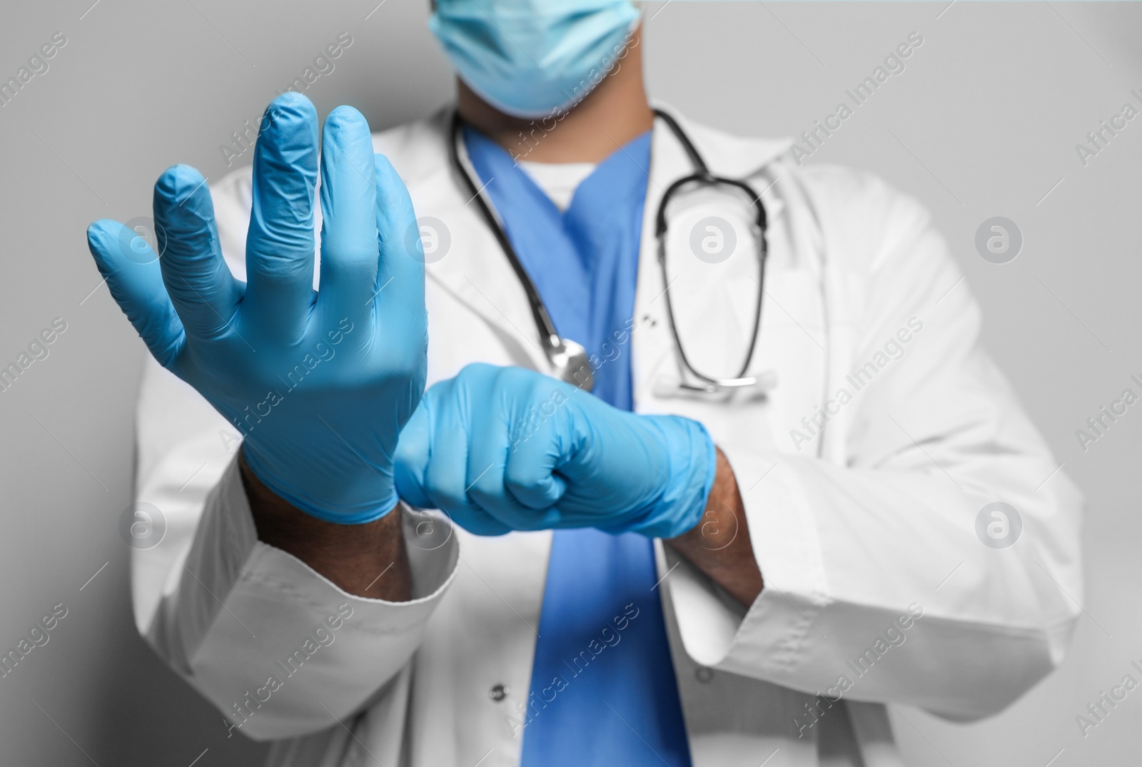 Photo of Doctor in protective mask putting on medical gloves against light grey background, closeup
