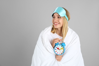 Young woman in sleeping mask wrapped with blanket holding alarm clock on grey background. Space for text