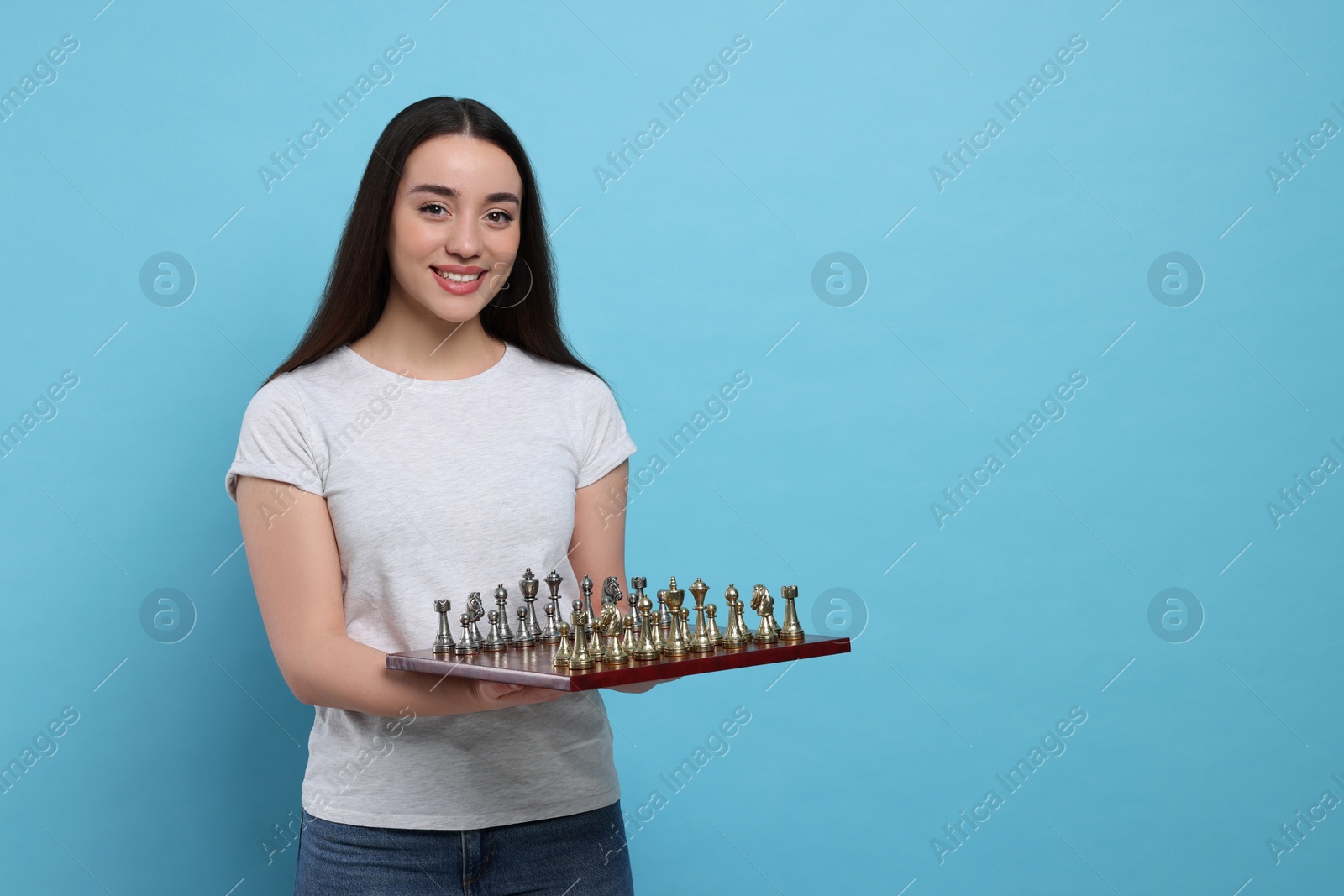 Photo of Happy woman holding chessboard with game pieces on light blue background, space for text