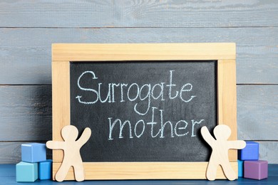 Photo of Small chalkboard with phrase Surrogate mother, people figures and cubes on blue wooden table