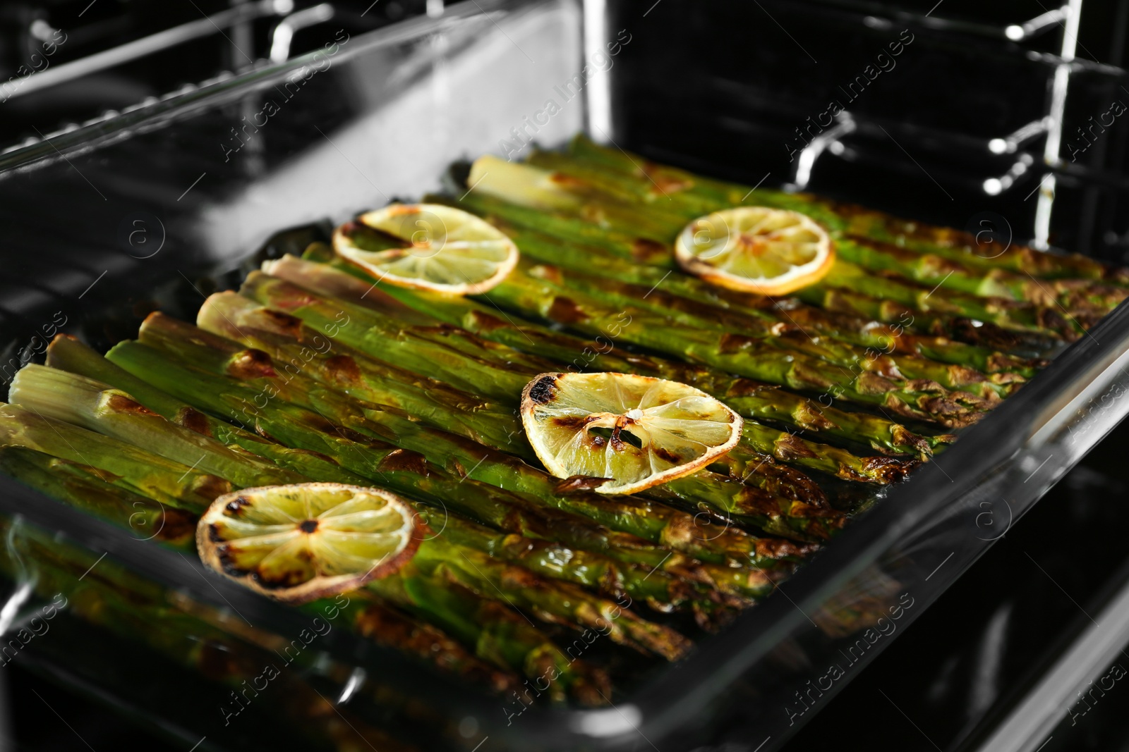 Photo of Cooked asparagus with lemon slices in glass baking dish on oven rack, closeup