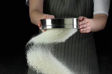 Photo of Woman sieving flour at table against black background, closeup