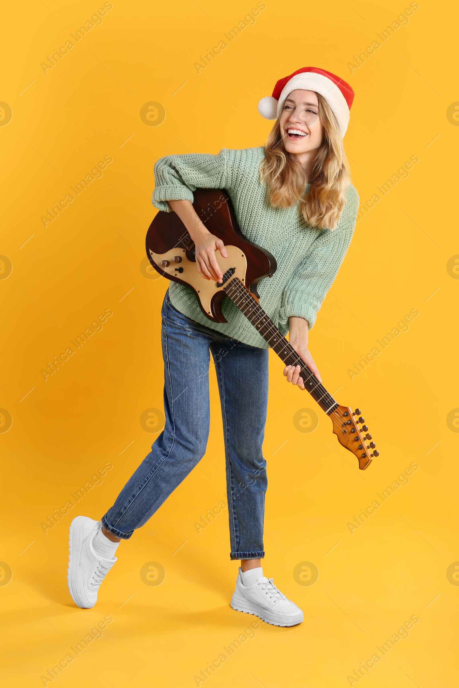 Photo of Young woman in Santa hat playing electric guitar on yellow background. Christmas music
