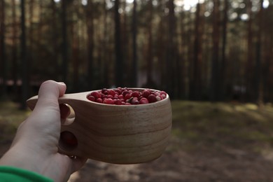 Photo of Woman holding wooden cup with tasty lingonberries near forest, closeup