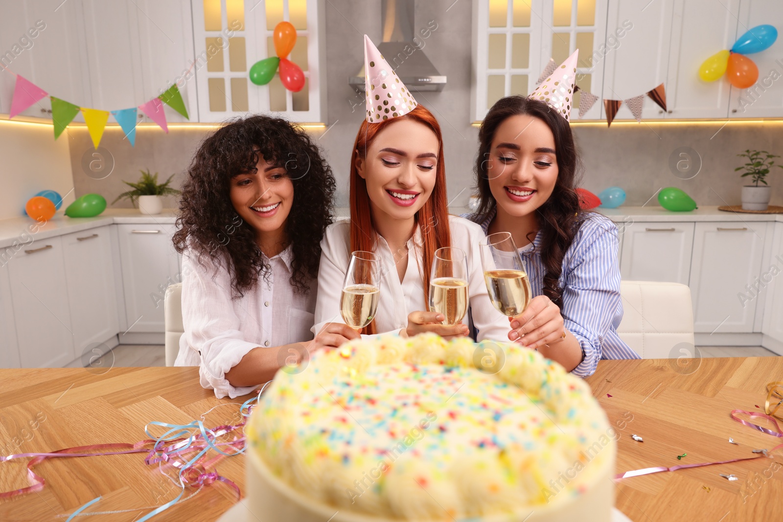 Photo of Happy young women with tasty cake and glasses of sparkling wine celebrating birthday in kitchen