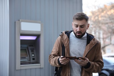 Photo of Unhappy young man with wallet near cash machine outdoors