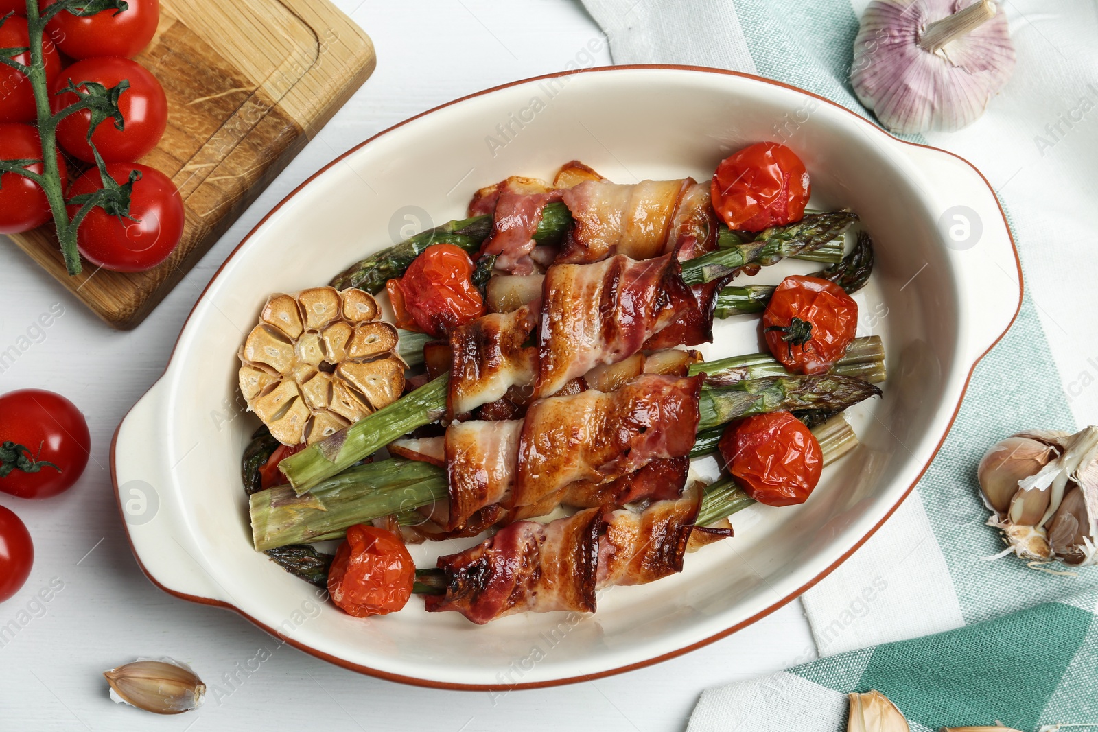 Photo of Oven baked asparagus wrapped with bacon in ceramic dish on white wooden table, flat lay