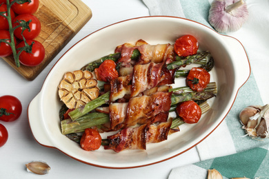 Photo of Oven baked asparagus wrapped with bacon in ceramic dish on white wooden table, flat lay