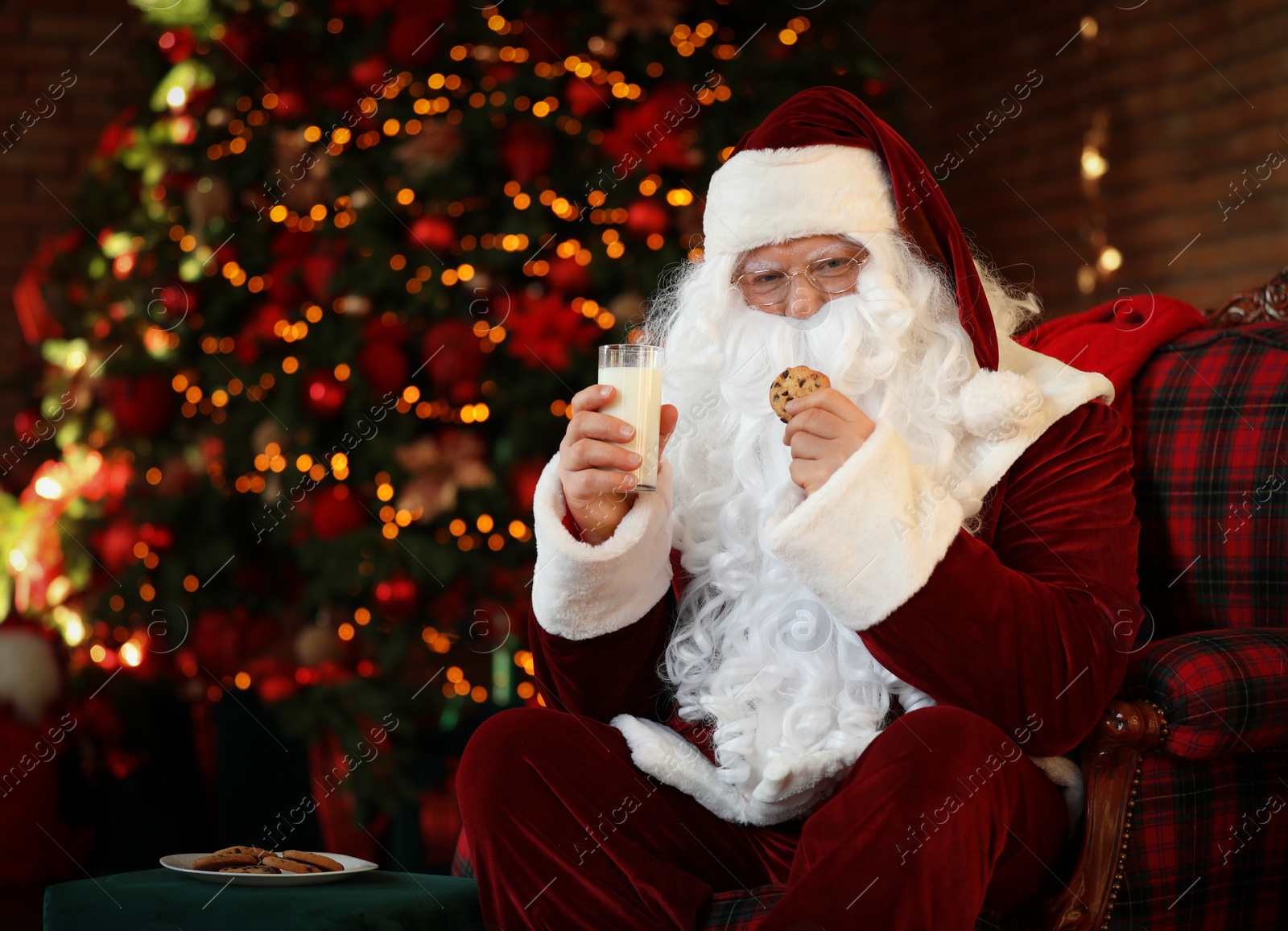 Photo of Santa Claus with milk and cookie near Christmas tree indoors