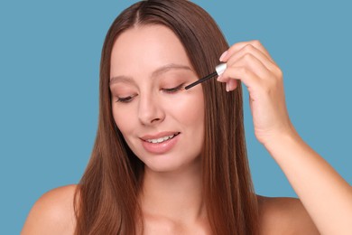 Beautiful woman applying serum onto eyelashes on light blue background