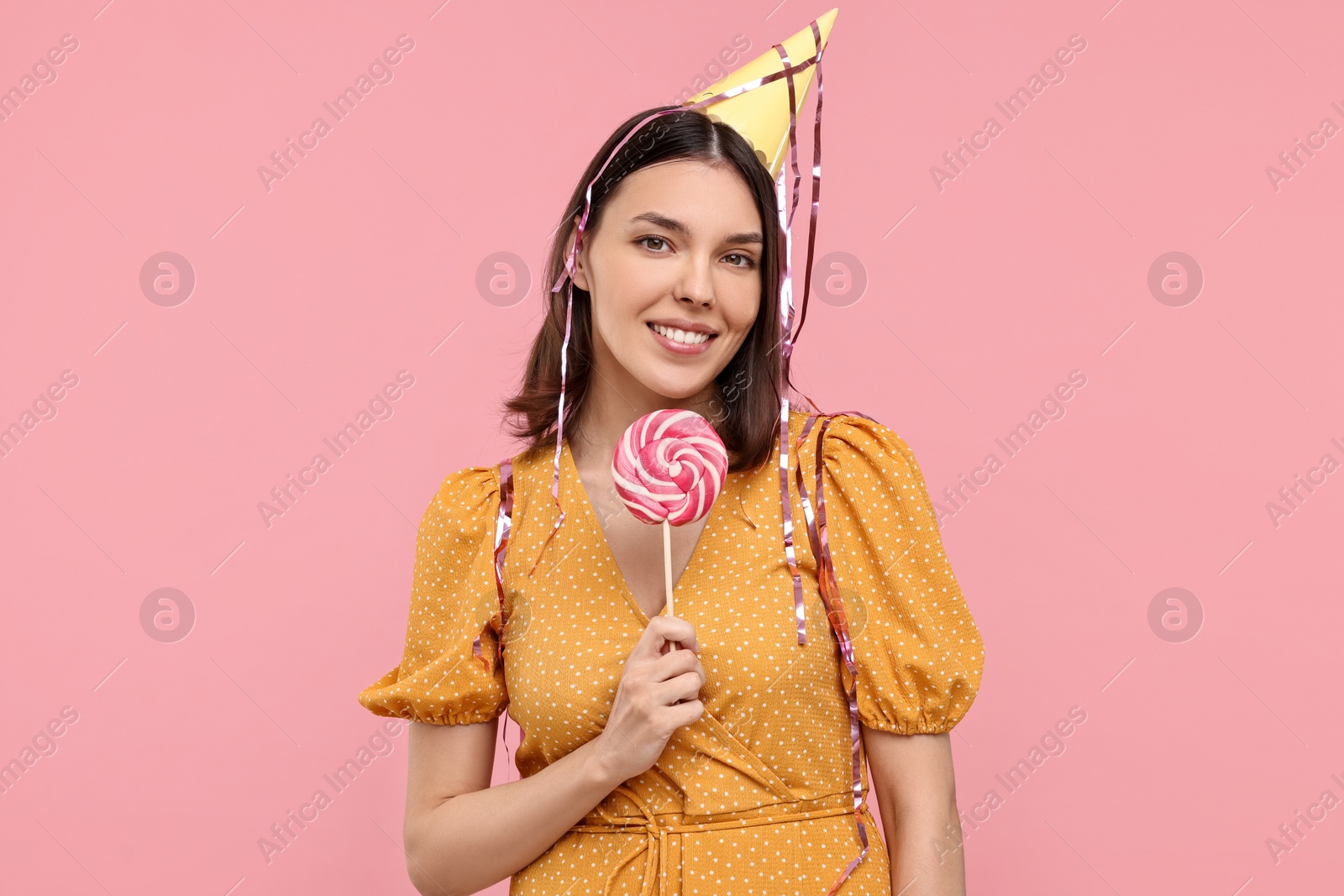 Photo of Happy young woman in party hat with candy on pink background