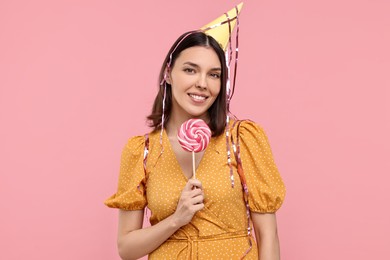 Happy young woman in party hat with candy on pink background