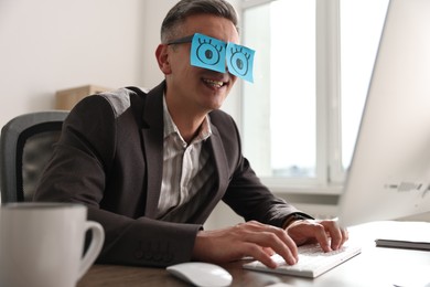 Man with fake eyes painted on sticky notes working on computer at table in office