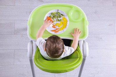Photo of Cute little baby eating healthy food in high chair indoors, top view