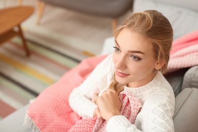 Beautiful young woman wrapped in plaid sitting on sofa at home. Winter atmosphere