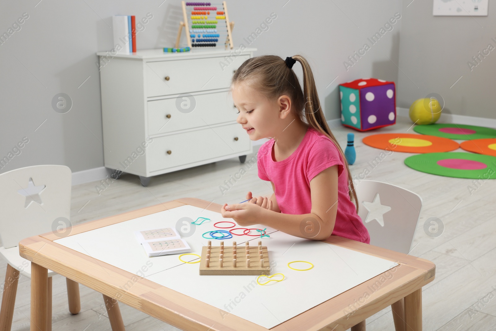 Photo of Motor skills development. Girl playing with geoboard and rubber bands at white table in kindergarten