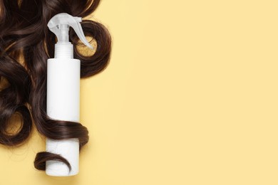 Spray bottle with thermal protection wrapped in lock of brown hair on yellow background, flat lay. Space for text