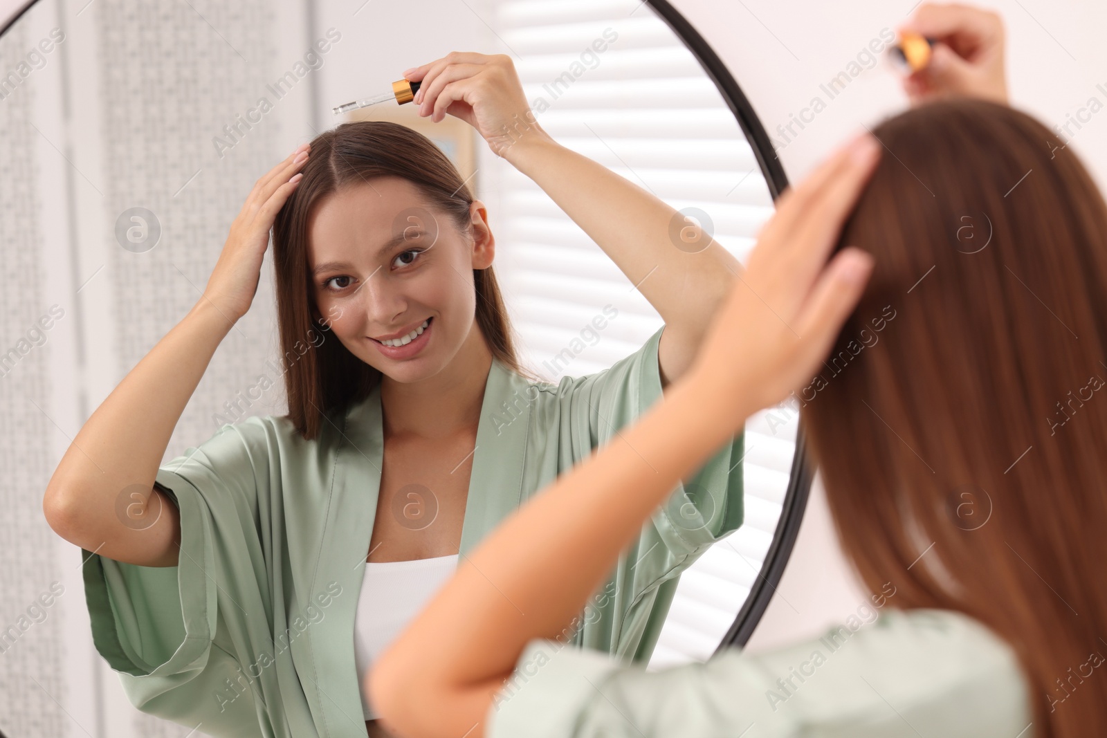 Photo of Beautiful woman applying serum onto hair near mirror indoors