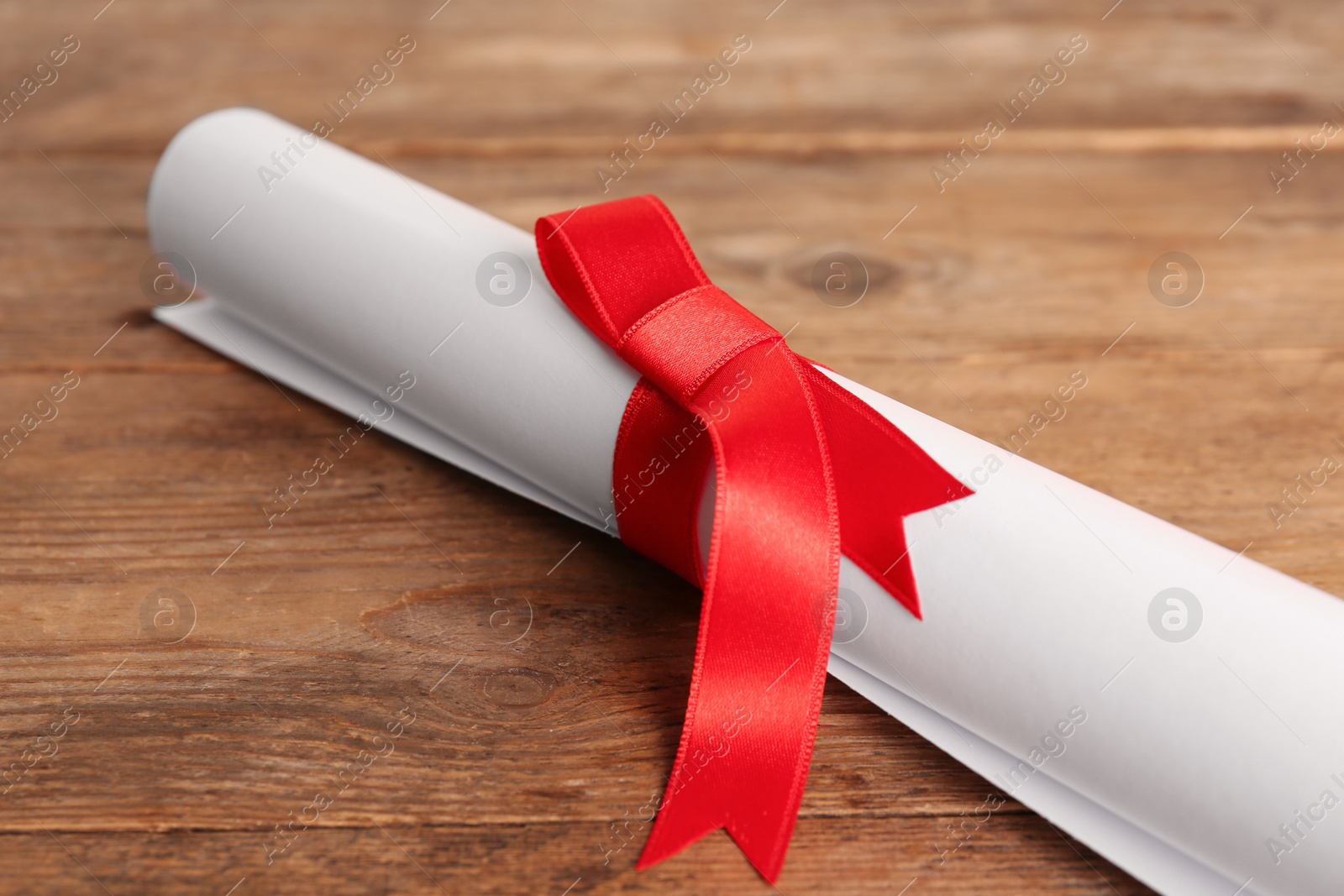 Photo of Rolled student's diploma with red ribbon on wooden table, closeup