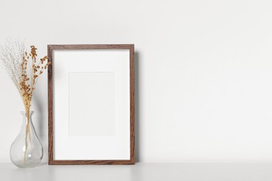 Empty photo frame and vase with dry decorative flowers on white table. Mockup for design