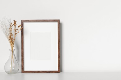 Photo of Empty photo frame and vase with dry decorative flowers on white table. Mockup for design