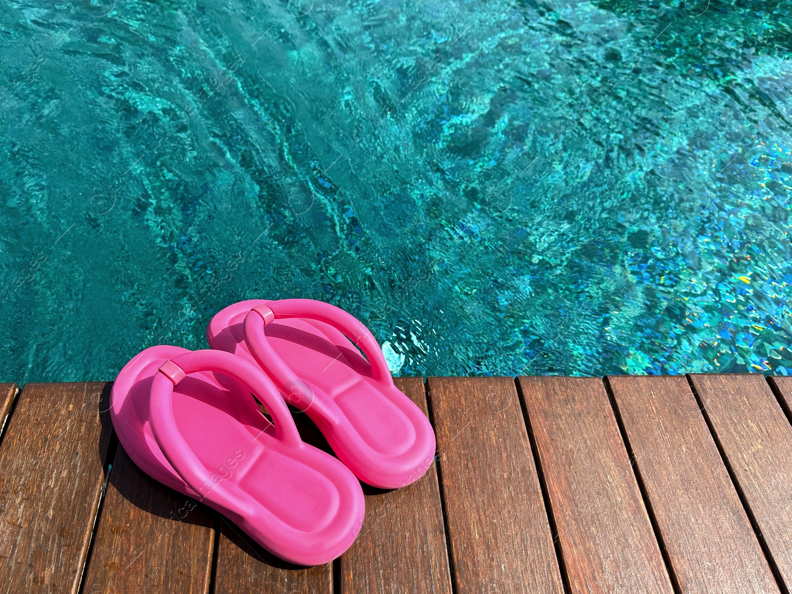 Photo of Clear rippled water in swimming pool and pink flip-flops on wooden deck outdoors. Space for text