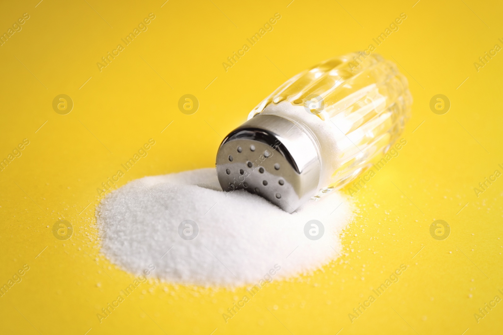 Photo of Scattered salt and shaker on yellow background, closeup