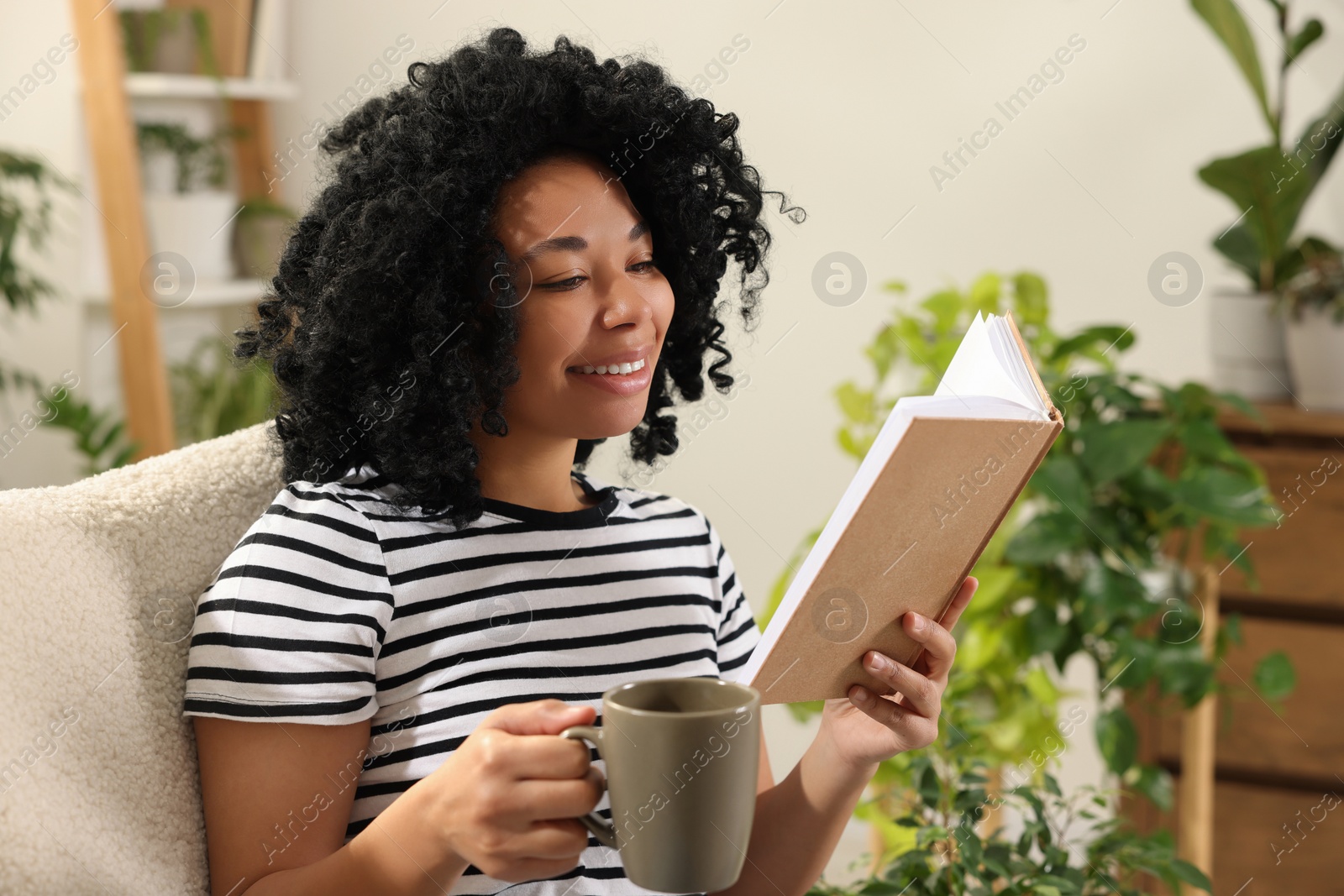 Photo of Relaxing atmosphere. Happy woman with cup of hot drink reading book surrounded by houseplants at home