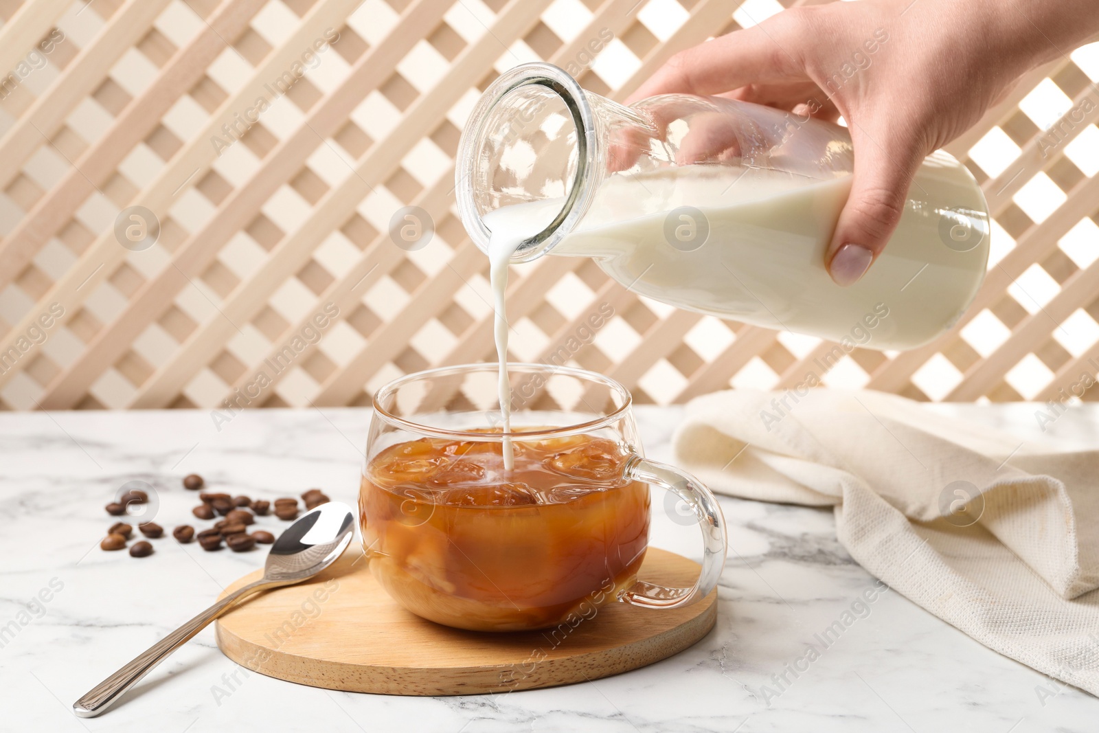 Photo of Woman pouring milk into glass with refreshing iced coffee at white marble table, closeup