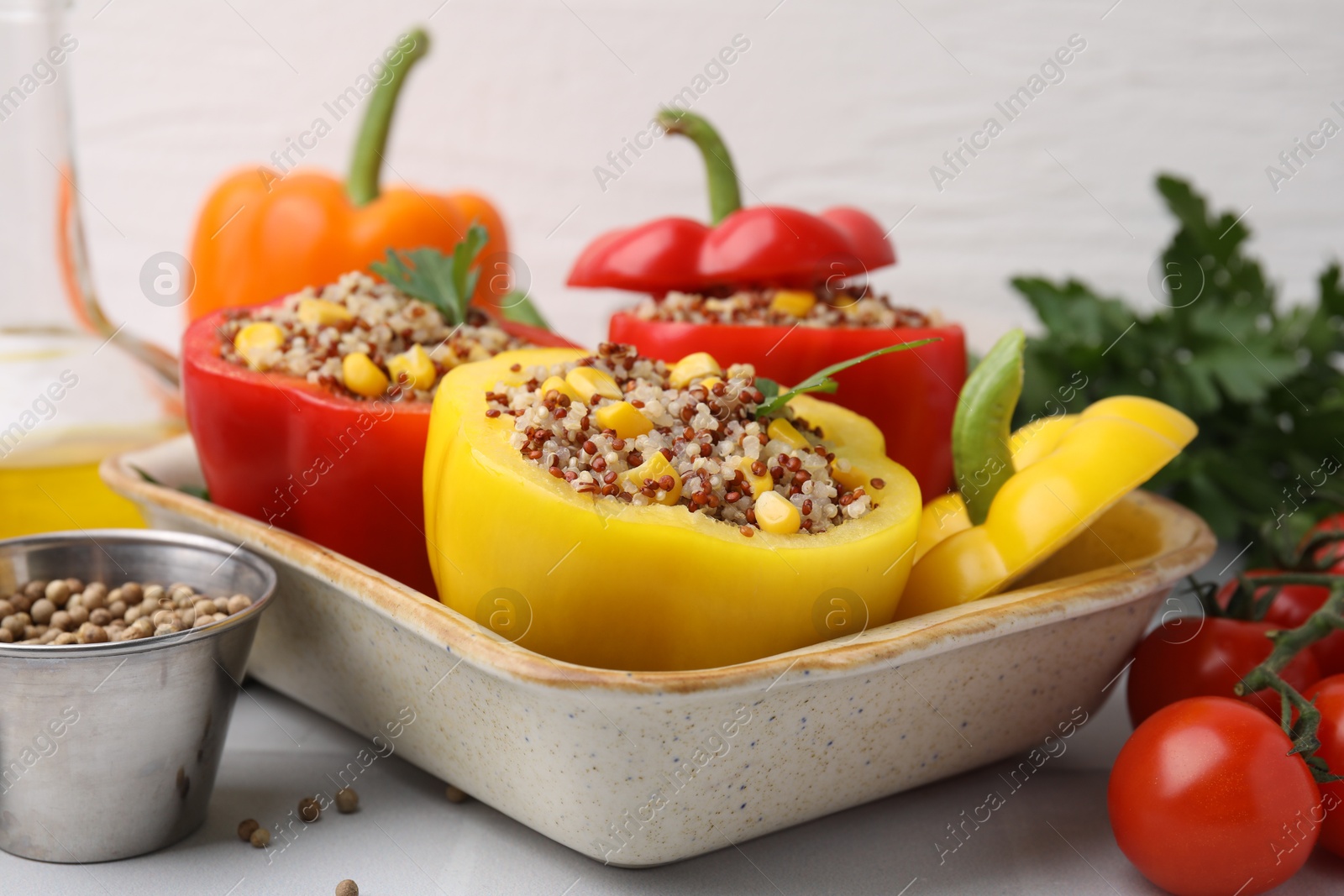 Photo of Quinoa stuffed bell peppers in baking dish, parsley, tomatoes and peppercorns on white tiled table, closeup