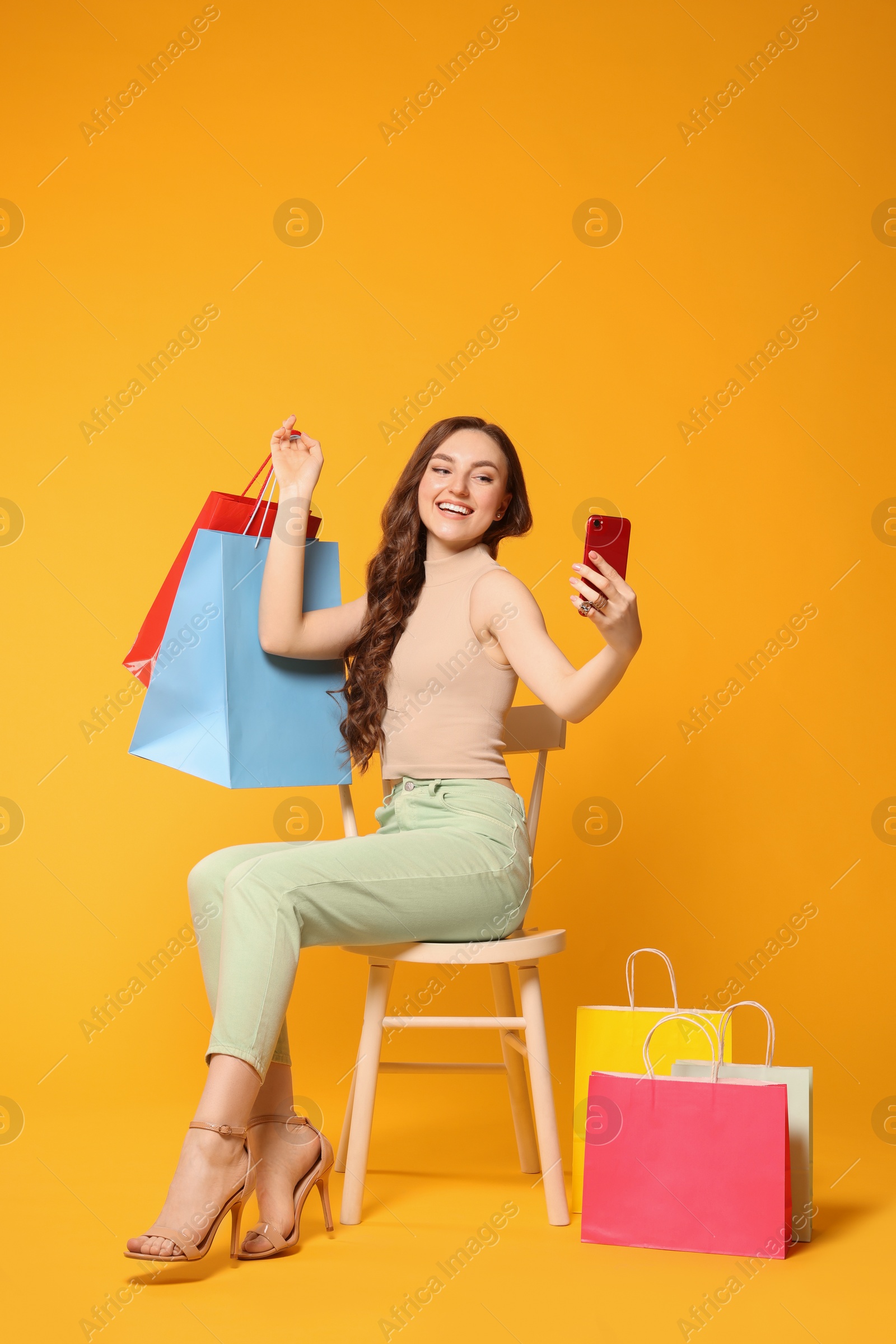 Photo of Beautiful woman taking selfie while holding colorful shopping bags on chair against orange background