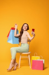 Beautiful woman taking selfie while holding colorful shopping bags on chair against orange background