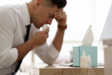 Photo of Ill businessman in office, focus on table with box of paper tissues, nasal spray and drops