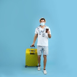 Male tourist in protective mask holding passport with ticket and suitcase on turquoise background