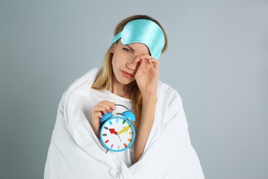 Young woman in sleeping mask wrapped with blanket holding alarm clock on grey background