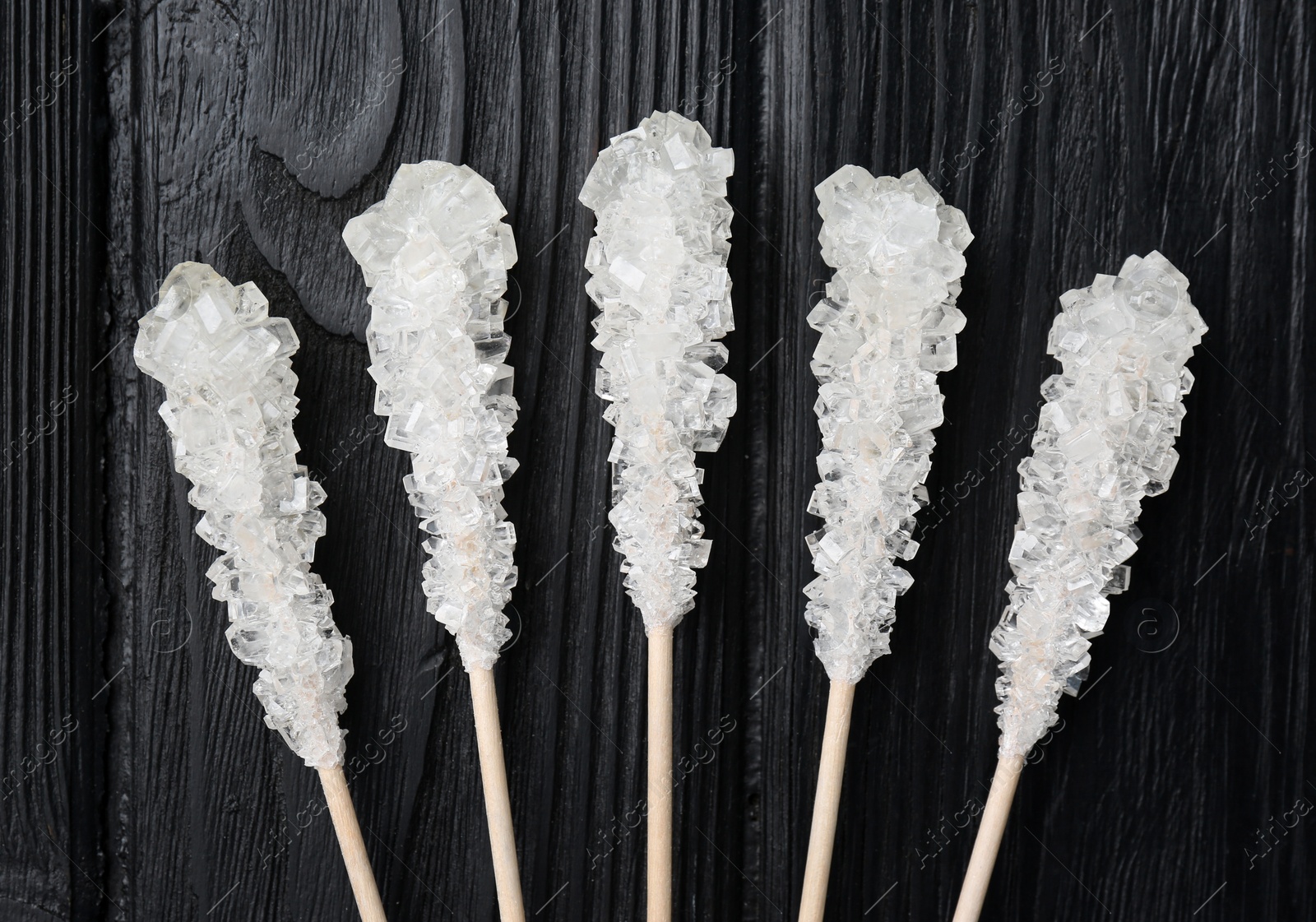 Photo of Sticks with sugar crystals on black wooden table, flat lay. Tasty rock candies